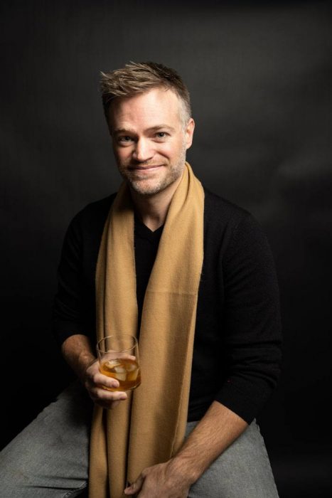 Casey sitting down in front of a black background with a tan scarf and holding a bourbon glass